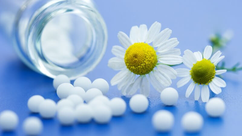 Homöopathie - Medikamentenglas und weiße Kügelchen mit zwei Gänseblümchen auf blauem Untergrund