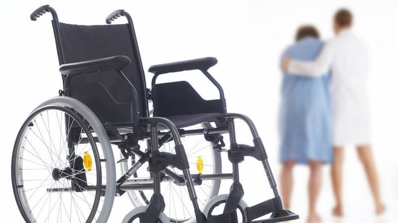 Rollstuhl, im Hintergrund hilft Ärztin einer Patientin beim Gehen