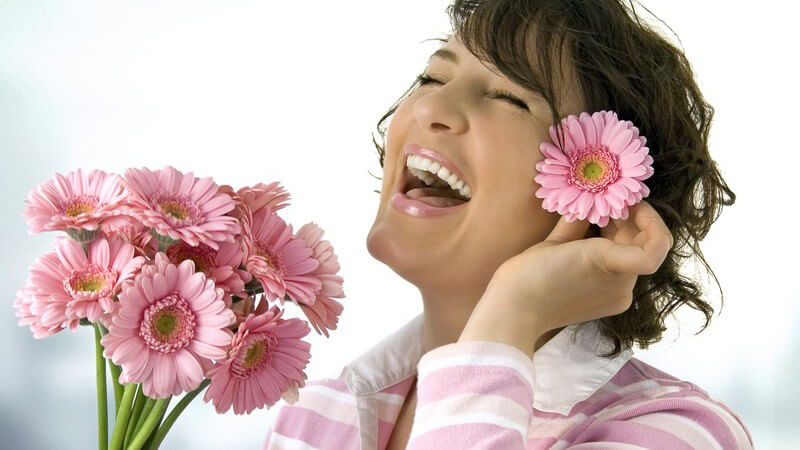 Lachende Frau hält rosanen Blumenstrauß in Hand, steckt sich eine Blüte hinters Ohr