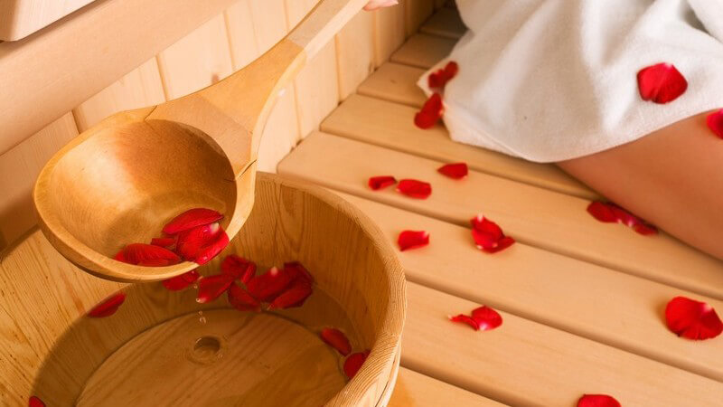 Holzeimer mit Wasser und Rosenblättern in Sauna neben Frau in weißem Handtuch
