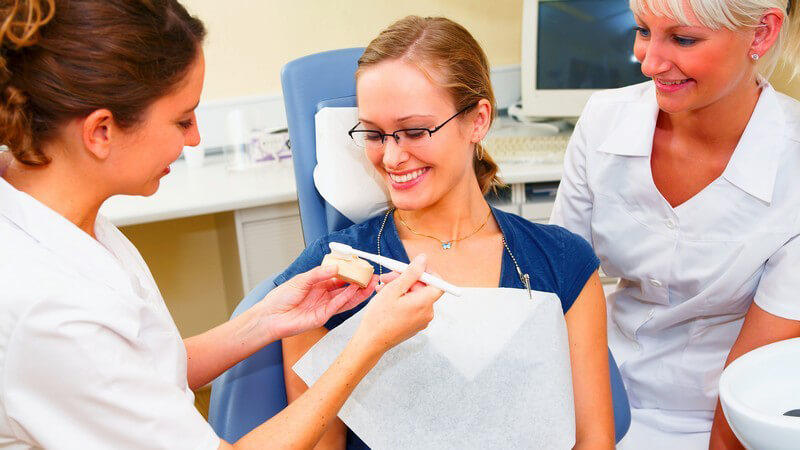Zwei Zahnarzthelferinnen sitzen in Praxis bei Patientin auf Zahnarztstuhl, zeigen das richtige Zähneputzen