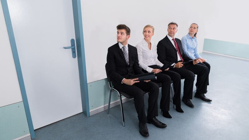 Zwei Männer im Anzug und zwei Frauen in Bluse sitzt wartend vor einem Büro im Flur
