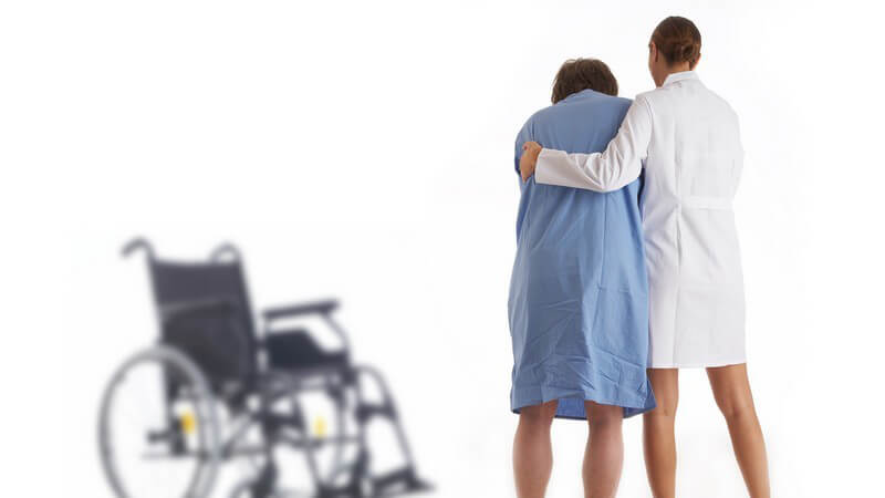 Rückenansicht, Krankenschwester stützt Patientin beim Gehen, im Hintergrund Rollstuhl