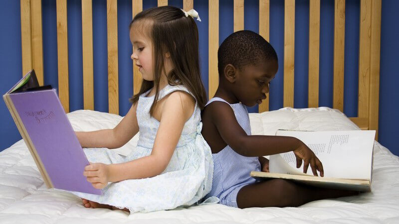 Zwei Kinder lesen in Büchern, ein hellhäutiges, brünettes Mädchen links und ein farbiger Junge in blauem Shirt rechts