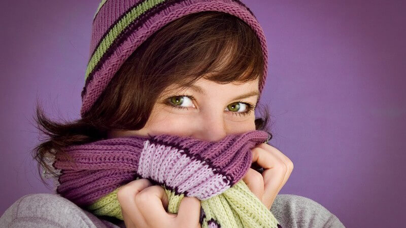 Junge Frau mit Wollmütze und Schal, versteckt Mund und Nase unter dem Schal, lächelt in Kamera