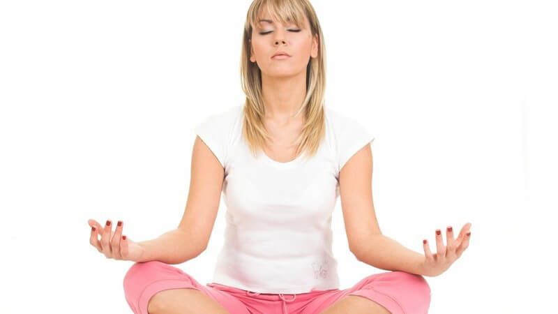 Blonde, junge Frau in rosa Sporthose und weißem Sport-Tshirt sitzt im Schneidersitz mit geschlossenen Augen und meditier
