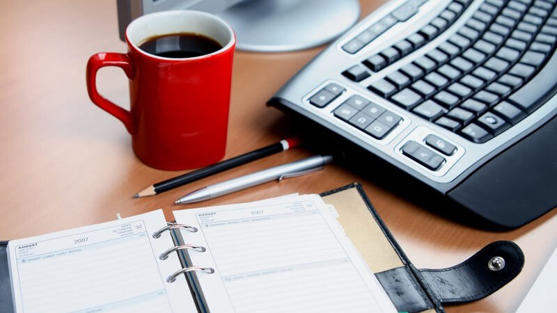 Schreibtisch mit PC, Terminkalender, Stiften und Becher Kaffee