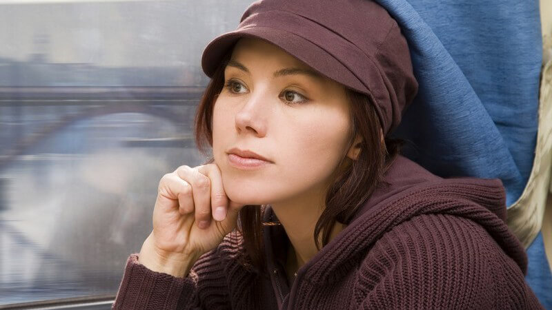 Brünette Frau mit brauner Mütze sitzt Verträumt am Zugfenster, im Hintergrund eine Brücke
