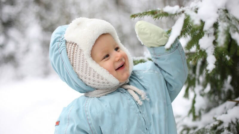 Kleinkind im Schneeanzug am Baum freut sich über Schnee