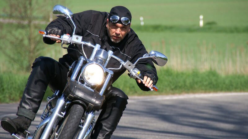 Motorradfahrer in schwarzer Kleidung, Kopftuch kommt um Kurve gefahren auf Kamera zu