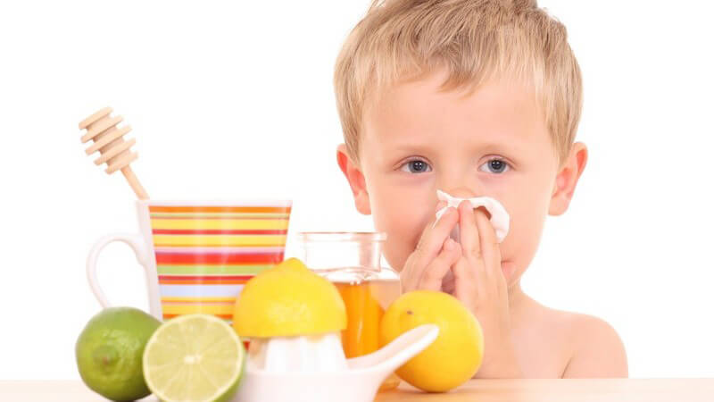 Kleiner Junge mit Erkältung putzt sich Nase, davor Tee, Limetten, Zitronen, Honig auf Tisch