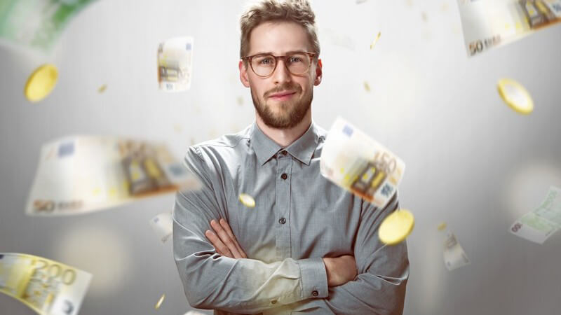 Junger Mann mit Brille und grauem Hemd posiert vor herunterregnenden Euro-Geldscheinen und -Münzen