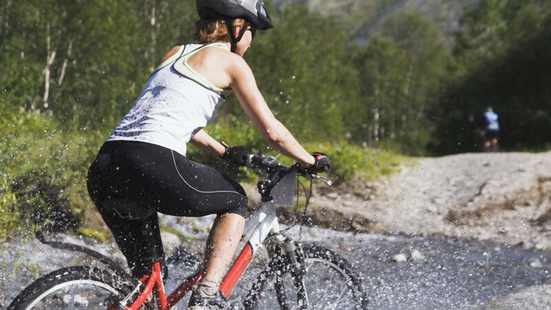 Frau fährt auf Mountainbike durch Wasser mit Helm und Sportbekleidung und Wald und Bergen im Hintergrund