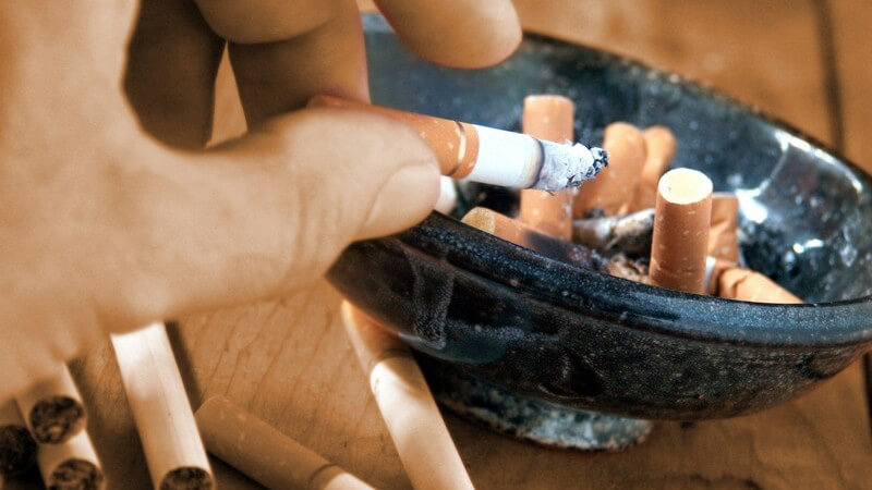 Nahaufnahme männliche Hand drückt Zigarette in vollem Aschenbecher aus, daneben weitere Zigaretten