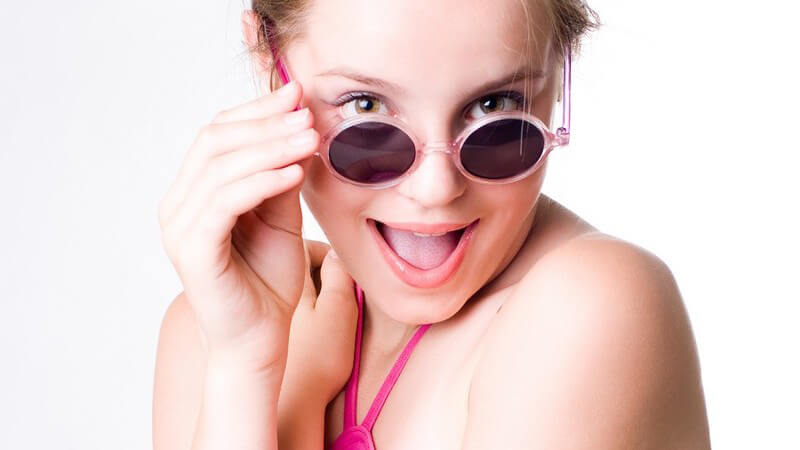 Junge Frau mit Sonnenbrille im Sommerkleid schaut frech mit offenem Mund in Kamera