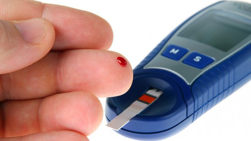 Linke Hand mit Blutstropfen auf Fingerkuppe neben einem Bluttest Gerät