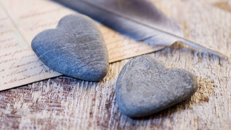 Nahaufnahme zwei Herzen aus Stein, im Hintergrund Postkarte, Schreib-Feder