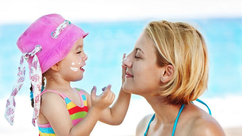 Mädchen mit pinkem Sonnenhut cremt ihrer Mutter am Strand das Gesicht ein