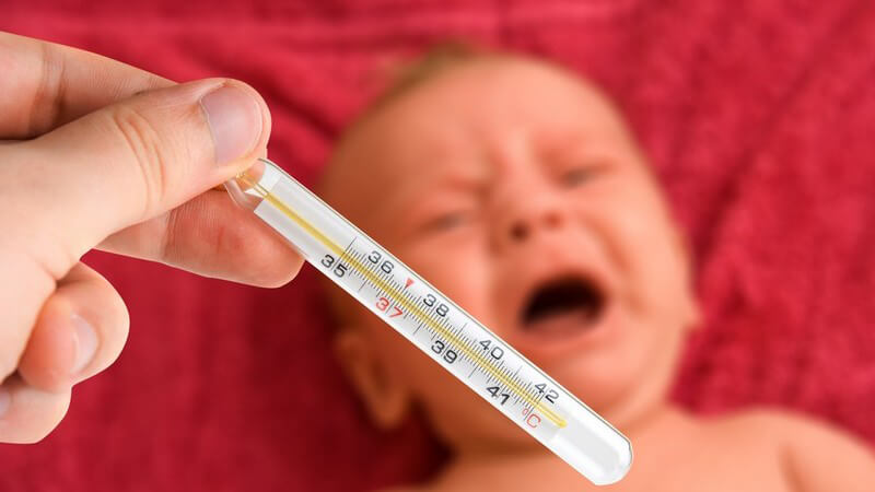 Hand hält Fieberthermometer, im Hintergrund schreiendes Baby