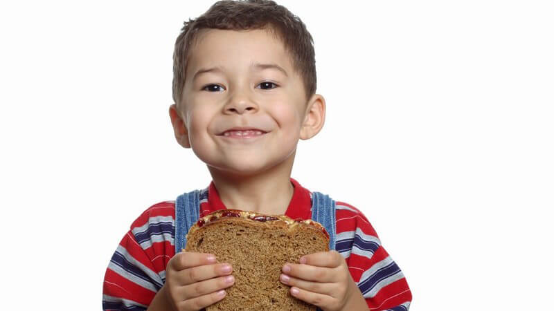 Kleiner grinsender Junge hält Brot mit Erdnussbutter zufrieden in Händen