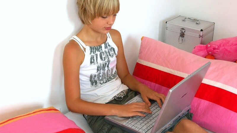 Blondes, kurzhaariges Mädchen neben rosa Kissen mit Laptop auf dem Schoß