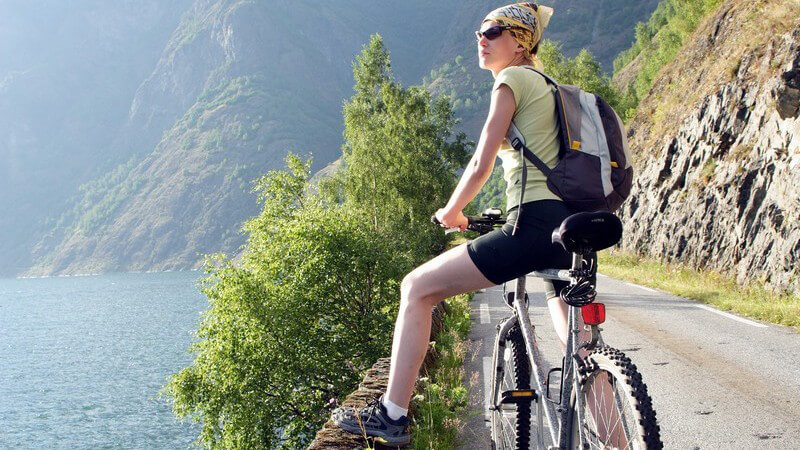 Frau auf Fahrrad oder Mountainbike an Bergstraße mit Sonnenbrille und Kopftuch, die Pause macht und auf Bergsee schaut