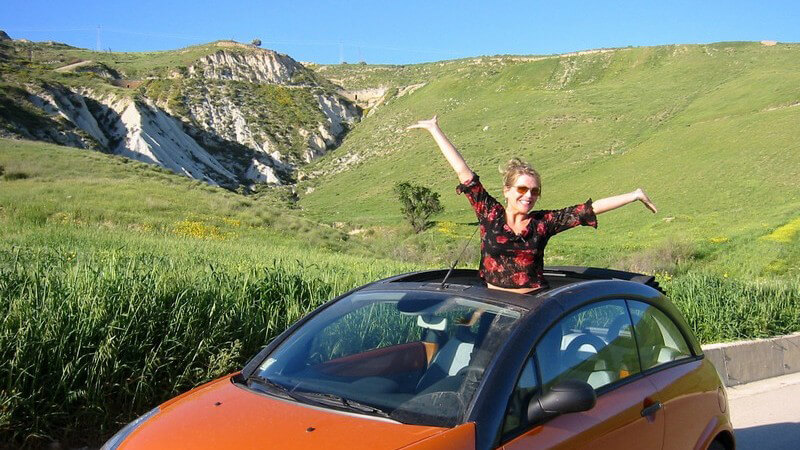 Frau steht im Auto und schaut aus Schiebedach hervor, im Hintergrund Berglandschaft