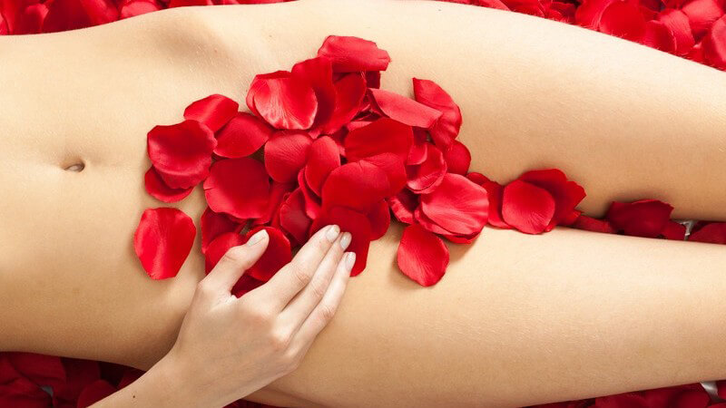Nackter Frauenkörper in einem Bett von roten Rosenblättern, der Intimbereich ist mit Rosenblättern bedeckt