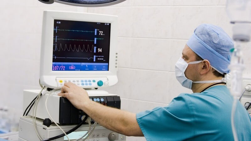 Mann in medizinischer Schutzkleidung kontrolliert mit der linken Hand einen Monitor mit medizinischen Messdaten