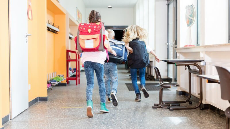 Drei Kinder mit Tornister auf dem Rücken laufen nach Schulende über den Flur in der Schule