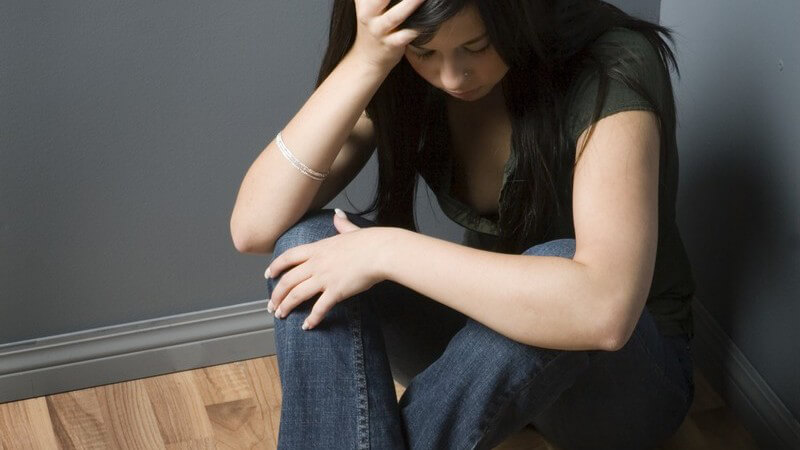Junge depressive Frau sitzt in Zimmerecke, neben ihr Tabletten auf Boden