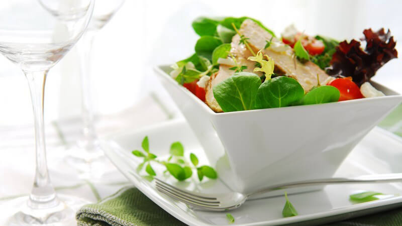 Salat mit Hähnchenstreifen in hoher, quadratischer Schale mit Unterteller und grüner Serviette