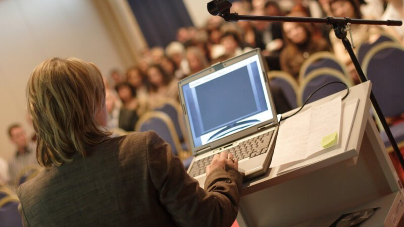 Frau steht mit Laptop vor Podium bei einem Vortrag, in Richtung Publikum fotografiert