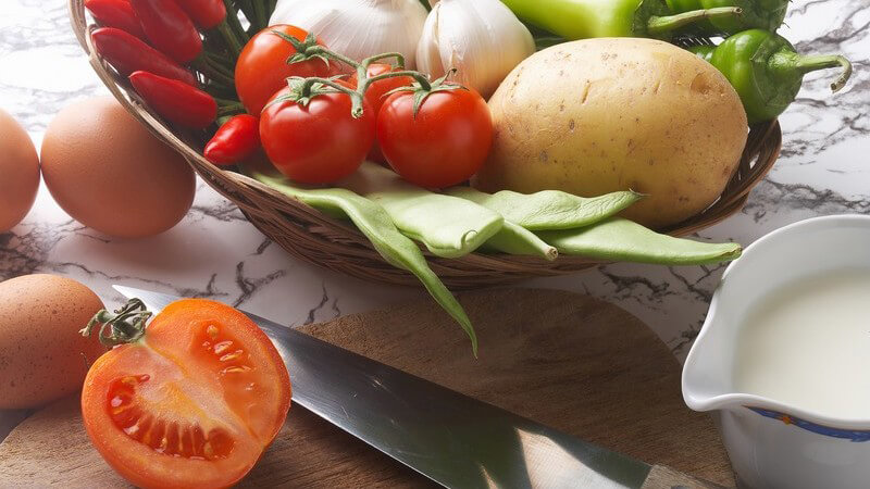 Zutaten - Messer und Tomatenhälfte auf Schneidebrett, daneben Eier, Milch, Chili, Tomaten, Knoblauch und eine Kartoffel