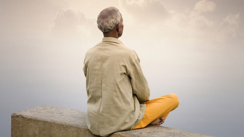 Ein älterer, indischer Mann in gelber Hose sitzt meditierend auf einer Mauer
