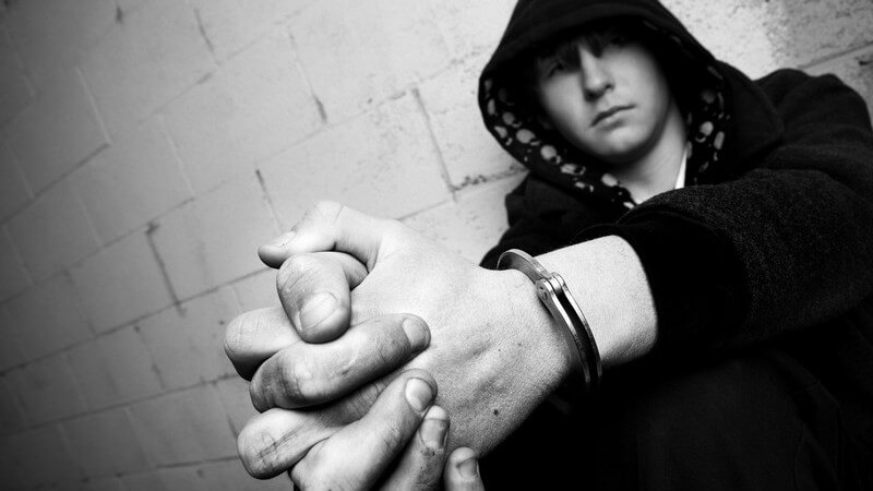 Schwarz-weiß Bild Jugendlicher in Kapuzenpulli hockt an Mauer, Hände in Handschellen