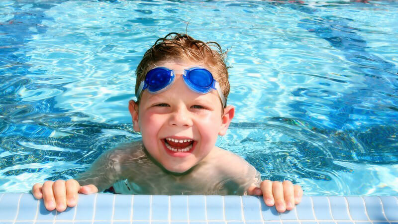 Kleiner Junge mit Schwimmbrille auf Stirn am Beckenrand im Schwimbad lacht, im Hintergrund rot-weiße Absperrleine