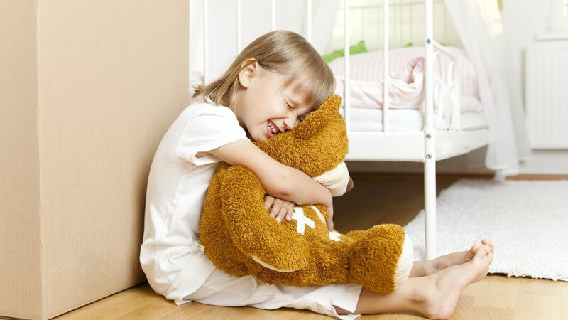 Kleines Mädchen sitzt vor ihrem Bett und hält Teddy fest im Arm