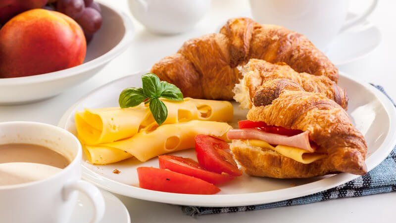 Brunch Frühstücksteller mit Croissants, Käse und Tomaten, daneben eine Kaffeetasse und ein Obstteller