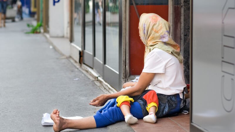 Bettlerin mit Kopftuch in Armut sitzt an Straßenrand mit Baby, bettelt um Geld, wendet Blick ab