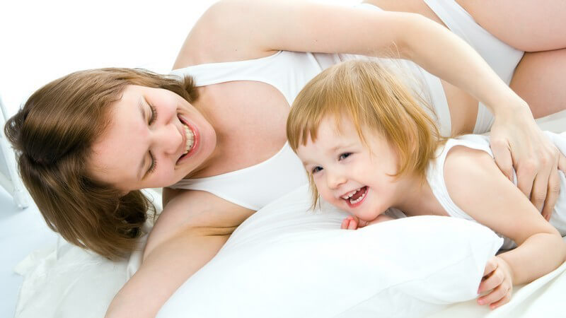 Mutter spielt lachend mit kleinem Mädchen, Tochter, in weißer Unterwäsche auf weißer Bettdecke