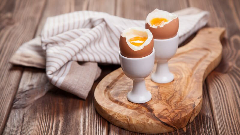 Zwei gekochte und geköpfte Eier im Eierbecher auf Holzbrett neben Küchenhandtuch