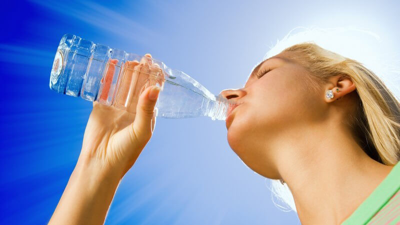 Junge, blonde Frau unter blauem Himmel trinkt Wasser aus einer Plastikflasche