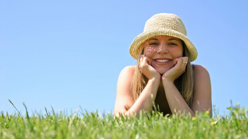 Mädchen mit Strohhut liegt lächelnd auf einer Wiese, den Kopf in die Hände gelegt