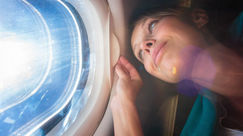 Junge Frau blickt glücklich durch ein Fenster im Flugzeug, die Sonne scheint hindurch