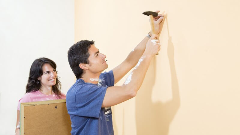 Paar beim Renovieren, Mann schlägt Nagel mit Hammer in Wand, Frau hält Bild zum Aufhängen