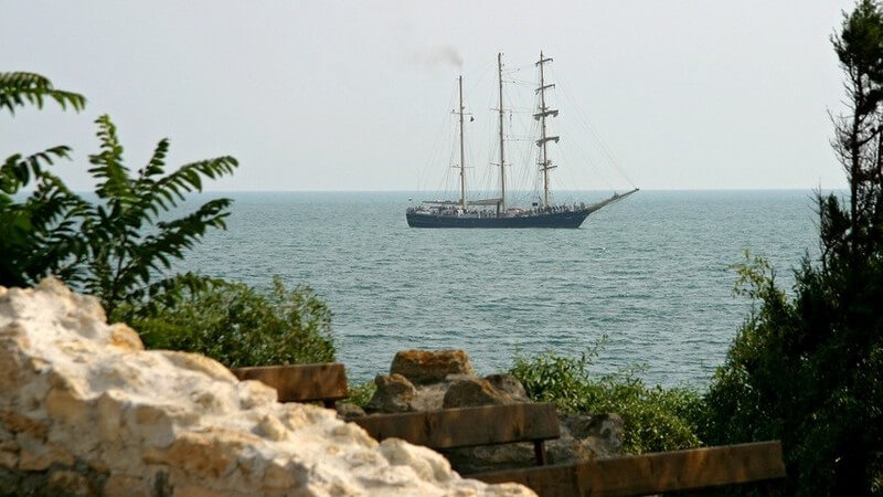 Segelschiff im Hintergrund mit eingeholten Segeln auf dem Schwarzen Meer, im Vordergrund Felsen und Sträucher