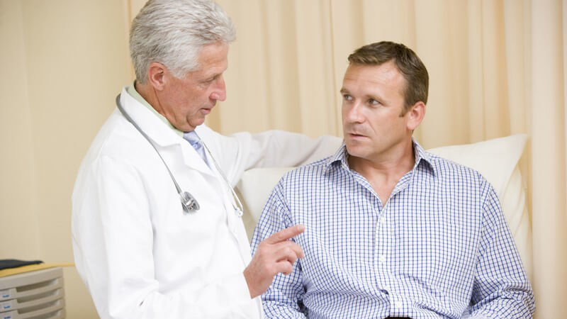 Ergrauter Arzt mit Stethoskop um den Hals redet mit Mann in blauem Hemd