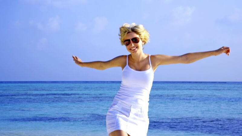 Junge Frau im Sommerkleid streckt Arme aus, im Hintergrund Meer