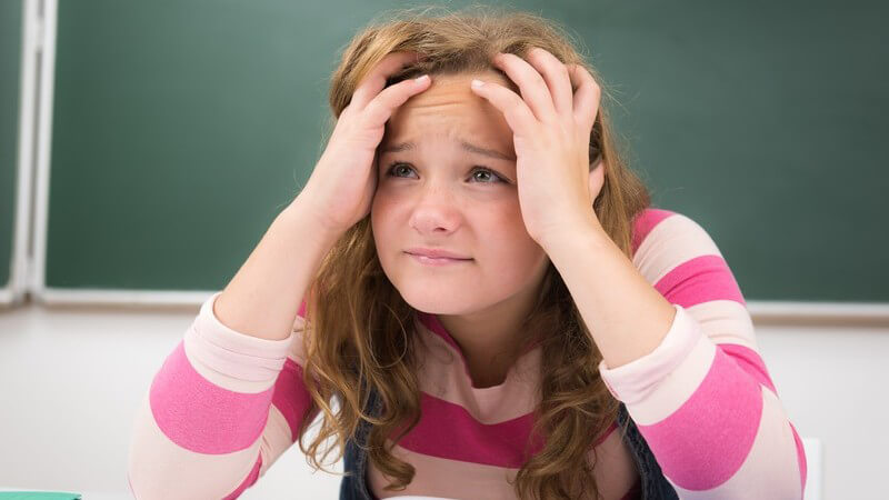 Mädchen sitzt über einem Heft im Klassenzimmer und fasst sich verzweifelt und überfordert an den Kopf
